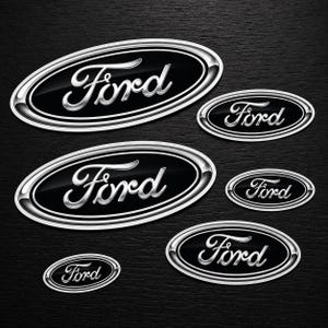 May include: A selection of Ford oval badges in different sizes. Each badge displays the word "Ford" in a silver script font on a black background, enclosed by a silver border. The badges are set against a dark, textured backdrop.