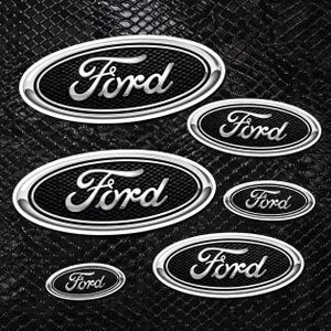 May include: A collection of Ford emblem decals in various sizes. Each oval emblem features the Ford script in silver against a black textured background, framed by a silver border. The decals are arranged on a black, textured surface.