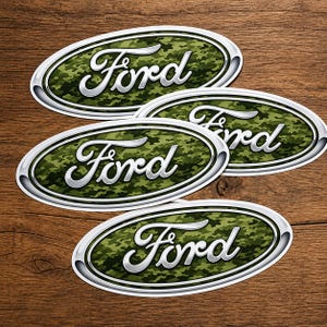 Könnte beinhalten: Vier ovale Ford-Embleme mit einem Tarnmuster in Grün- und Brauntönen. Das Wort "Ford" ist in silberner Schreibschrift geschrieben. Jedes Emblem hat einen silbernen Rand. Auf einem Holzgrund.