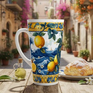 Può includere: Tazza alta in ceramica bianca con un manico grande, con un design a limone e onde in blu, giallo e bianco. La tazza è su un tavolo di legno con una piccola bottiglia di olio e una pasticceria.
