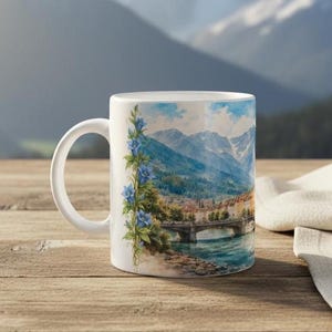 Könnte beinhalten: Weiße Keramiktasse mit einem malerischen Aquarellgemälde einer Berglandschaft, Brücke und Fluss. Die Tasse hat einen gebogenen Henkel und ist mit blauen Blumen verziert. Die Tasse steht auf einer Holzoberfläche.