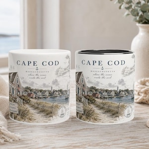 Könnte beinhalten: Zwei weiße Keramikbecher mit einem Cape Cod-Design. Die Becher zeigen eine Küstenlandschaft mit Häusern, einem Strand und dem Text "Cape Cod Massachusetts where the ocean meets the soul". Ein Becher hat eine schwarze Innenseite.