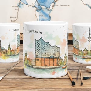 Op de afbeelding: Drie witte keramische mokken met een aquarel illustratie van Hamburg, Duitsland. Elke mok toont een ander aanzicht van de skyline van de stad, met het woord "Hamburg" erboven. De mokken staan op een houten oppervlak.