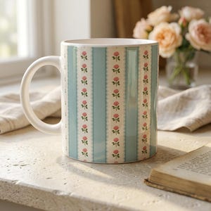Könnte beinhalten: Eine weiße Keramiktasse mit einem hellblau-weißen Streifenmuster. Die Streifen sind mit kleinen rosa Rosen-Illustrationen verziert. Die Tasse hat einen weißen Henkel und steht auf einer hellen Oberfläche.