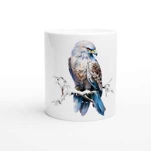 Könnte beinhalten: Eine weiße Keramiktasse mit einer bunten Illustration eines Habichts, der auf einem schneebedeckten Ast sitzt. Der Habicht hat braune, graue und blaue Federn.