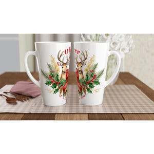 May include: Two white ceramic latte mugs with a Christmas theme. Each mug features a deer illustration with a red bow, holly, and the text "Oh Deer, it's Christmas time!" The mugs are on a wooden table with a checkered tablecloth.