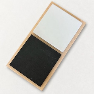 May include: A rectangular wooden base featuring two square tiles mounted on its surface. One tile is solid white with a smooth finish, while the other tile is solid black with a textured, matte appearance. The wooden frame has a light tan color.