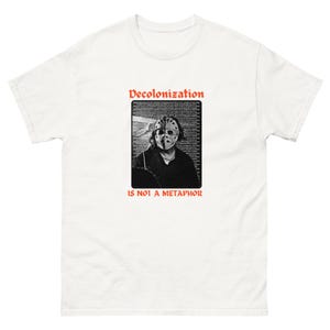 May include: A white short-sleeved t-shirt featuring a central graphic print. The design includes the red text Decolonization is not a metaphor and a black and white image of a person wearing a hockey mask over a background of dense text.