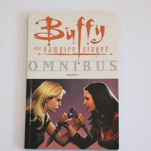 Puede incluir: Una portada de cómic con el título "Buffy Cazavampiros Omnibus Volumen 5". El arte de la portada representa a dos mujeres enfrentadas, una con cabello rubio y la otra con cabello oscuro, ambas sosteniendo armas.