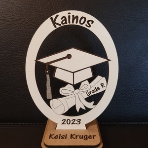 Op de afbeelding: Wit houten afstudeercadeau met een afstudeerpet, diploma en strik. Het cadeau is gegraveerd met de tekst "Kainos", "Grade R", "2023" en "Kelsi Kruger".