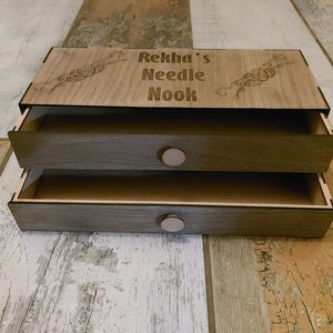 Puede incluir: Una caja de madera con dos cajones, cada uno con una perilla redonda. La parte superior de la caja está grabada con el texto "Rekha's Needle Nook" y un diseño de ovillos de lana.