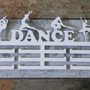Può includere: Espositore per medaglie in legno bianco con la parola "DANCE" ritagliata e quattro silhouette di ballerini. L'espositore ha sei slot per le medaglie.