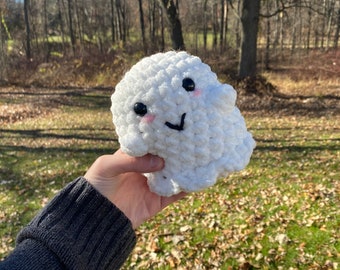 May include: A white crocheted ghost with black eyes and a pink blush. The ghost has a black smile and is being held in a hand.
