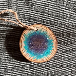 Può includere: Un ornamento rotondo in legno con un design a galassia dipinta. Il centro è viola scuro, circondato da un anello turchese con macchie bianche. Un anello di spago naturale è attaccato per appenderlo. L'ornamento misura circa 7.6 cm di diametro.