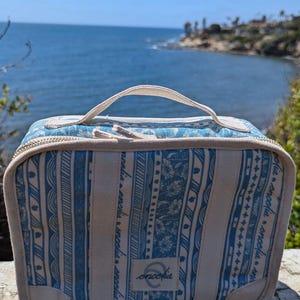 May include: A white and blue striped canvas zippered pouch with a brown leather handle. The pouch has a white label with the word "encolpia" printed on it. The pouch is sitting on a wooden surface.