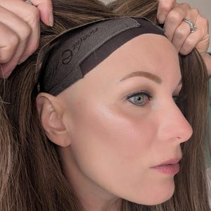 May include: A brown wig with highlights is held up, revealing a dark brown band with the brand name "neelia" printed on it. The wig is being held near a person's face, showing the hairline and the wig's construction.