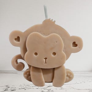 May include: A light tan monkey-shaped candle with a single white wick at the top. The candle features a stylized face with two circular eyes, a small nose, and a mouth. Each ear is decorated with a small heart-shaped cutout, and a curled tail.