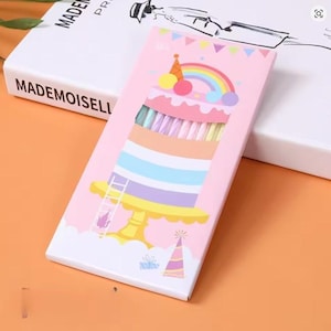 Può includere: Una scatola rosa con un design a torta, un arcobaleno, una scala e un cappello da festa. La scatola contiene oggetti colorati, forse matite o pennarelli. La scatola è su una superficie arancione, con un libro bianco sullo sfondo.