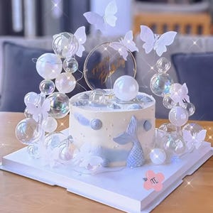 May include: A round white cake decorated with a blue mermaid tail, seashells, and small white pearls. The cake is surrounded by clear iridescent spheres and white butterfly cutouts. A circular gold-rimmed topper reads Happy Birthday.