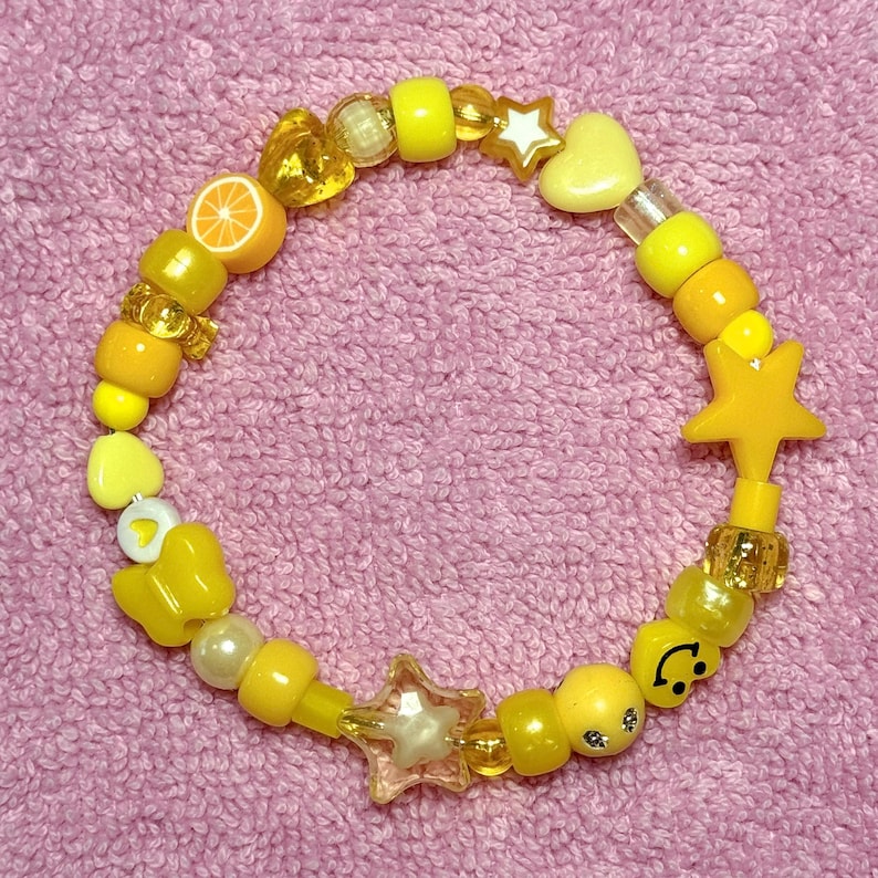 May include: A yellow and white beaded bracelet with a lemon slice, stars, hearts, and a smiley face charm.