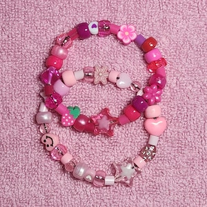 May include: Two pink and white beaded bracelets with various charms, including hearts, stars, flowers, and a strawberry. The bracelets are on a pink fuzzy surface.