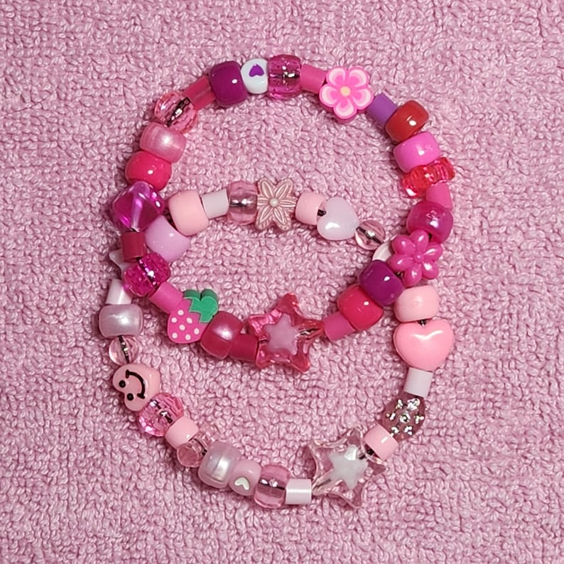 May include: Two pink and white beaded bracelets with various charms, including hearts, stars, flowers, and a strawberry. The bracelets are on a pink fuzzy surface.