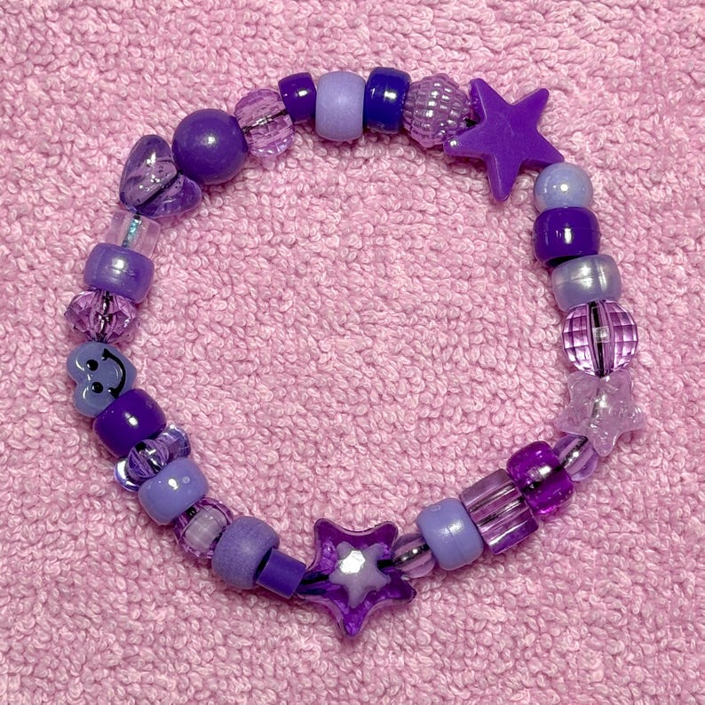 May include: Purple beaded bracelet with star and heart charms. The bracelet is made with various shades of purple beads, including clear, iridescent, and opaque.