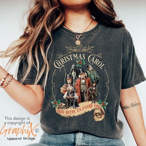 May include: A dark gray t-shirt with a vintage-style design featuring the characters from "A Christmas Carol". The design includes the text "Christmas Carol" in a decorative font, along with the quote "God Bless Us Every One".