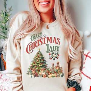 May include: A cream-colored sweatshirt with a vintage-style graphic of a Christmas tree, presents, and ornaments. The text "Crazy Christmas Lady" is printed in red and green.