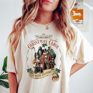 May include: A cream colored t-shirt with a vintage-style illustration of the ghosts from A Christmas Carol. The text reads "Christmas Carol" and "God Bless Us Every One".