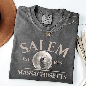 May include: A gray t-shirt with a vintage-style graphic design featuring the text "SALEM EST. 1626 MASSACHUSETTS" and a silhouette of a witch standing in front of a full moon.