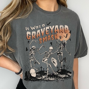 May include: Gray t-shirt with a graphic of three skeletons dancing in a graveyard. The text "It Was a Graveyard Smash" is printed on the shirt.