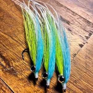 Puede incluir: Tres señuelos de pesca con largas fibras sintéticas fluidas en verde lima brillante, azul cielo y blanco. Cada señuelo tiene un ojo reflectante plateado y negro prominente unido a una cabeza envuelta en blanco y un anzuelo de metal negro curvo.