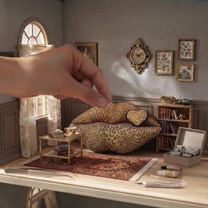 May include: A miniature dollhouse living room featuring a leopard print lip-shaped sofa with a matching heart-shaped pillow, a gold-toned coffee table, a patterned area rug, a wooden bookshelf, wall art, and a decorative gold wall clock.