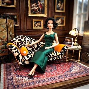 May include: A doll with dark hair styled in a vintage updo, wearing a floor-length emerald green satin gown. The doll sits on a sofa upholstered in a black and white leopard print fabric with orange accents, holding a small orange circular accessory.