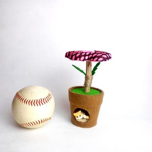 May include: A baseball next to a whimsical felt flower pot with a pink and black patterned top. The flower pot has a small opening with a miniature cat figurine inside. The flower stem is wrapped in twine.