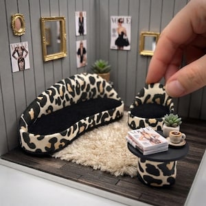May include: A miniature living room set featuring a curved leopard print sofa with black upholstery, a matching leopard print armchair, and a round black side table. The set includes a small white rug, a tiny potted succulent, and magazines.