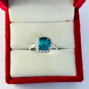 May include: A ring with a large, square-shaped blue gemstone surrounded by a halo of smaller, clear gemstones. The ring is set in silver and is displayed in a red velvet ring box.