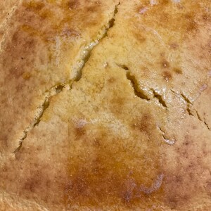 May include: A close-up of a golden brown cornbread with a crack down the center. The cornbread is slightly glossy and appears to be freshly baked.
