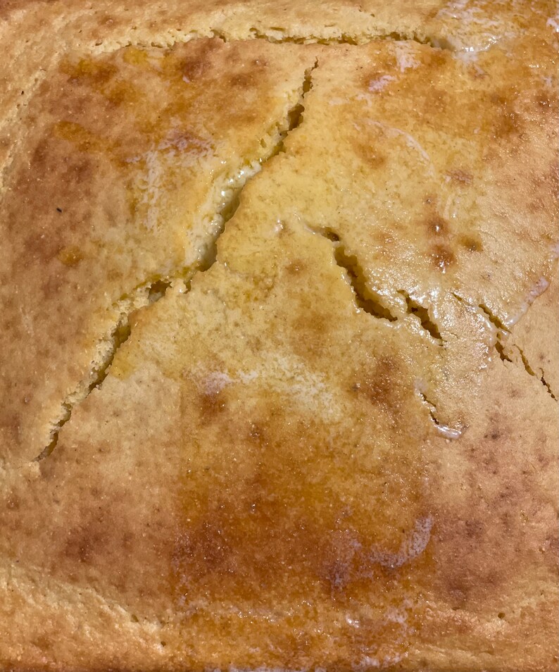 May include: A close-up of a golden brown cornbread with a crack down the center. The cornbread is slightly glossy and appears to be freshly baked.