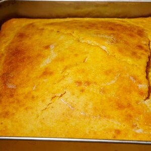 May include: A golden yellow cornbread baked in a square baking pan. The cornbread has a slightly cracked surface.