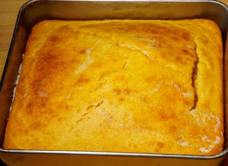 May include: A golden yellow cornbread baked in a square baking pan. The cornbread has a slightly cracked surface.