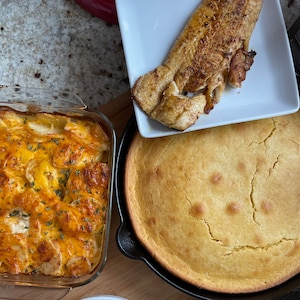 May include: A close-up of a casserole dish filled with a cheesy potato casserole, a skillet with a golden brown cornbread, and a white plate with a piece of fried fish.