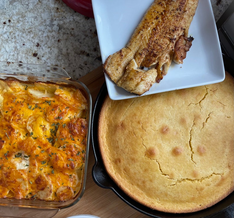May include: A close-up of a casserole dish filled with a cheesy potato casserole, a skillet with a golden brown cornbread, and a white plate with a piece of fried fish.