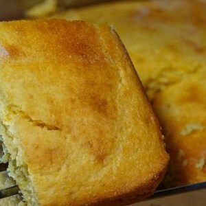 May include: A close-up of a golden yellow cornbread loaf being lifted from a baking pan with a metal spatula. The cornbread is soft and fluffy, with a slightly crispy crust.
