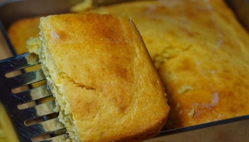 May include: A close-up of a golden yellow cornbread loaf being lifted from a baking pan with a metal spatula. The cornbread is soft and fluffy, with a slightly crispy crust.