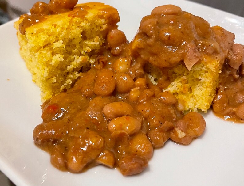 May include: A white plate with two pieces of cornbread and a brown gravy with white beans.