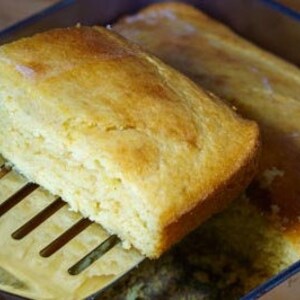 May include: A close-up of a golden yellow cornbread loaf being lifted out of a glass baking dish with a metal spatula. The cornbread is soft and fluffy, with a slightly crispy crust.