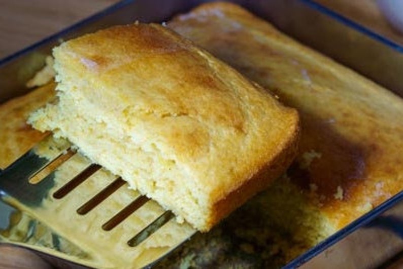 May include: A close-up of a golden yellow cornbread loaf being lifted out of a glass baking dish with a metal spatula. The cornbread is soft and fluffy, with a slightly crispy crust.