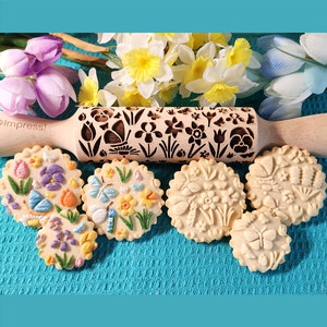 May include: A wooden rolling pin with a floral design, used to create embossed patterns on cookies. The rolling pin is surrounded by five cookies with different floral designs, including butterflies and flowers.